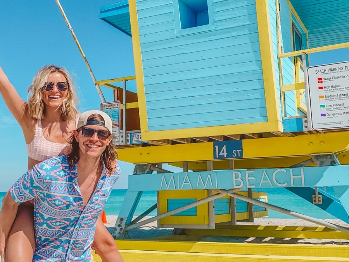 A couple is having fun at Miami Beach near a colorful lifeguard station. One person is giving a piggyback ride, both smiling and enjoying the sunny day.