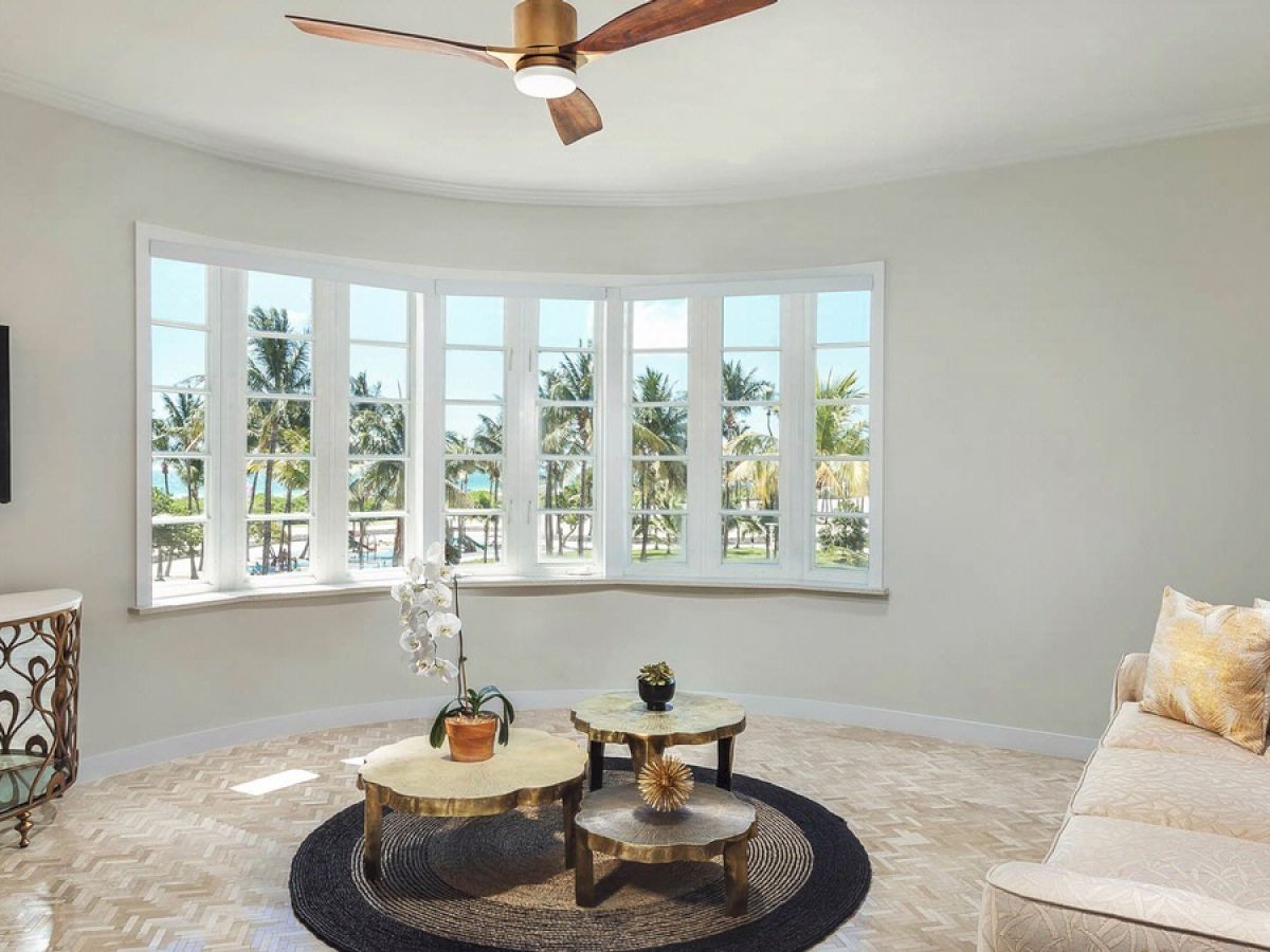 A spacious living room with a large window offering a scenic view of palm trees, sofa, coffee table, wall-mounted TV, and ceiling fan is depicted.