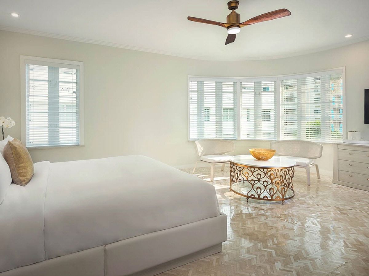 A modern bedroom features a large bed, white furniture, a decorative coffee table, wall-mounted TV, and ample natural light from multiple windows.