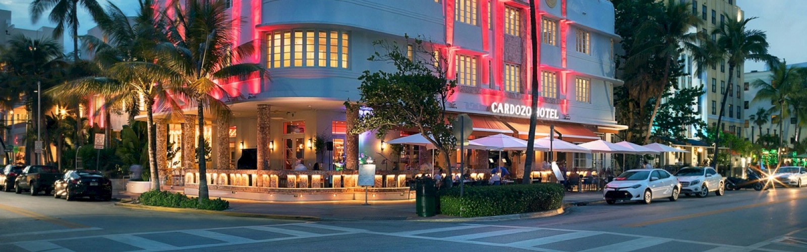 The image shows a vibrant street corner with the Cardozo Hotel, featuring bright lighting, palm trees, parked cars, and outdoor dining areas.