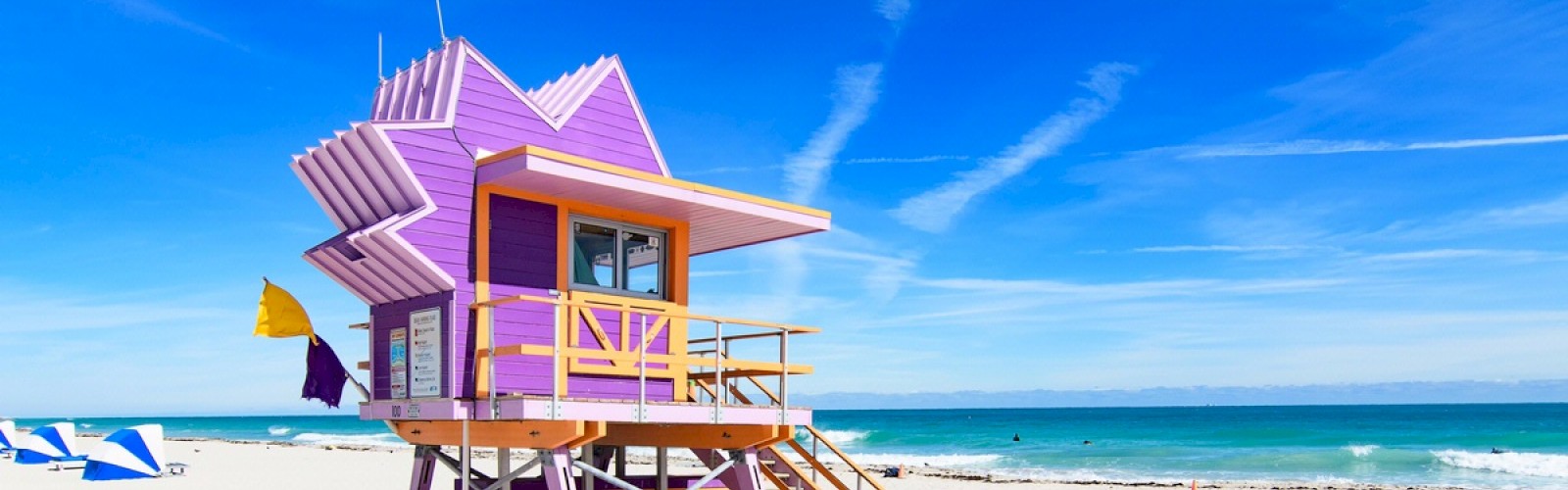 A vibrant purple lifeguard tower stands on a sandy beach against a clear blue sky, with the ocean waves gently hitting the shore in the background.
