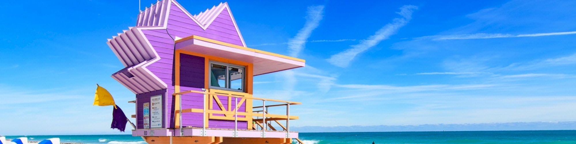 A vibrant purple lifeguard tower stands on a sandy beach against a clear blue sky, with the ocean waves gently hitting the shore in the background.