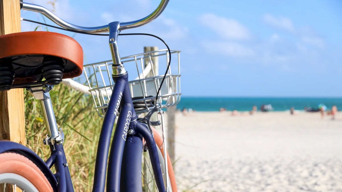 The image features a parked bicycle with a basket at a sandy beach, with people in the distance and a clear blue sky.