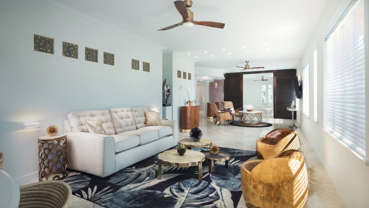 A modern living room featuring a white couch, wooden coffee tables, patterned rug, ceiling fan, and large windows with blinds, creating a cozy ambiance.