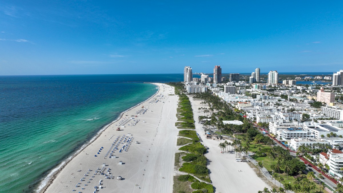 Aerial view of a beachfront city with sandy shores, turquoise waters, beach umbrellas, and high-rise buildings along the coast ending the sentence.