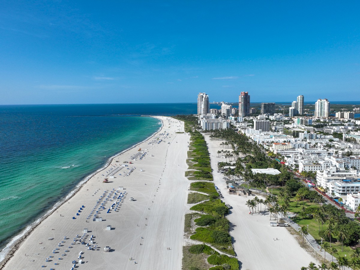 Aerial view of a beachfront city with sandy shores, turquoise waters, beach umbrellas, and high-rise buildings along the coast ending the sentence.