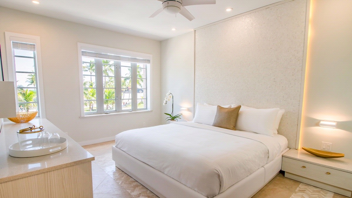 The image shows a modern bedroom with a large bed, white bedding, a ceiling fan, and natural light from windows. There's a dresser and a small plant.