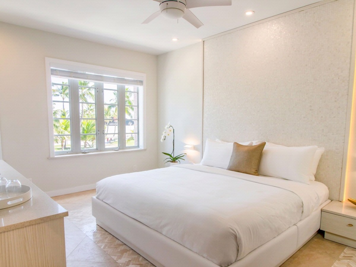 The image shows a modern bedroom with a large bed, white bedding, a ceiling fan, and natural light from windows. There's a dresser and a small plant.