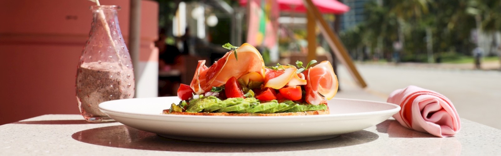 A plate of food is on a table outdoors, featuring avocado toast with toppings. Pink umbrellas and a beachside setting are in the background.