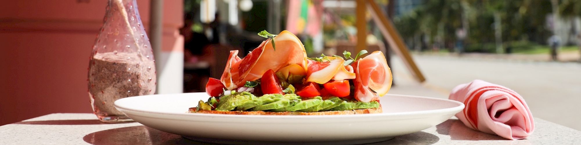 A plate of food is on a table outdoors, featuring avocado toast with toppings. Pink umbrellas and a beachside setting are in the background.