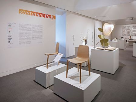 The image shows an exhibition with modern chairs on pedestals, an informative text on the wall, and various contemporary art pieces on display.