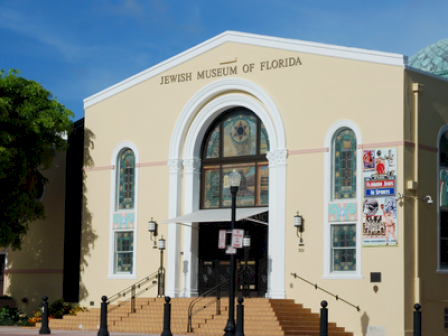 The image shows the front entrance of the Jewish Museum of Florida, with steps leading up and banners displayed on the right side of the building.