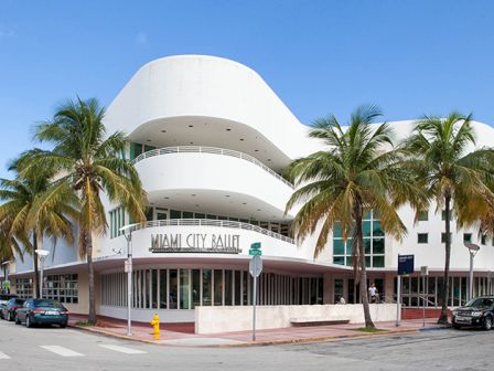 The image shows the Miami City Ballet building, a white, modern structure with curved architecture, surrounded by palm trees and parked cars.