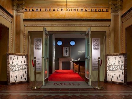 The image shows the entrance to Miami Beach Cinematheque, featuring posters for a Chaplin event, red carpet, and display cases inside.