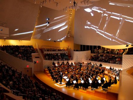 This image shows an orchestra performing on stage in a concert hall, with a seated audience watching and large projections on the walls.