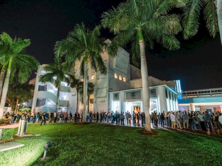 A large crowd is gathered in front of a building surrounded by palm trees at night, possibly for an event or opening.
