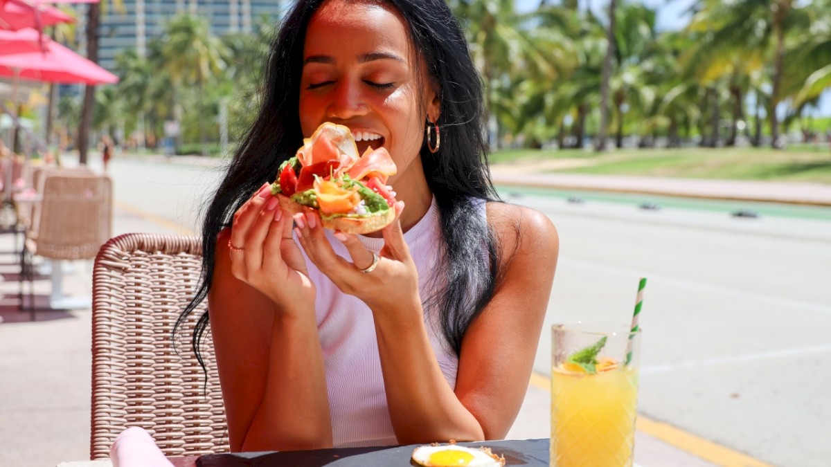 A woman is enjoying a meal outdoors at a sunny location with tropical palm trees and a drink beside her plate.