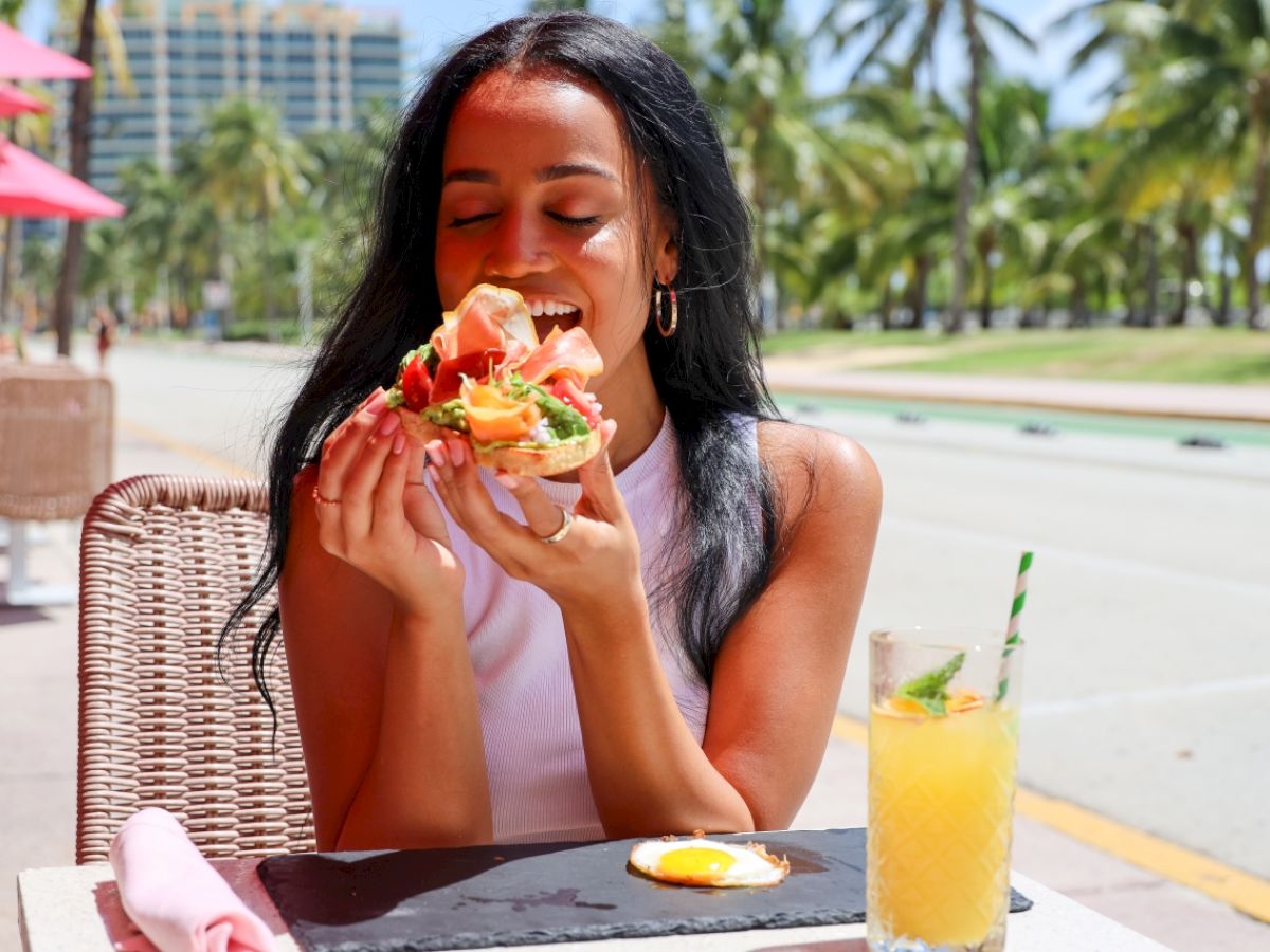 A woman is enjoying a meal outdoors at a sunny location with tropical palm trees and a drink beside her plate.