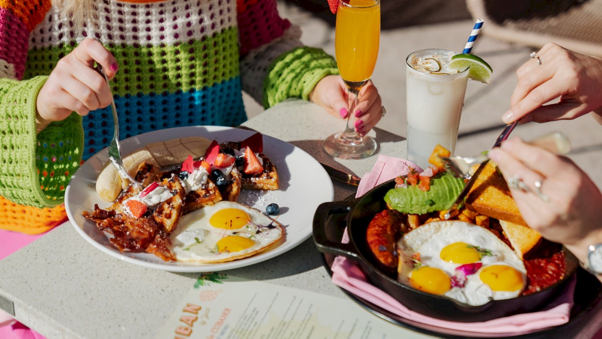 Two people enjoying a meal outdoors with plates of breakfast food, including eggs, waffles, and beverages on a table with a menu.