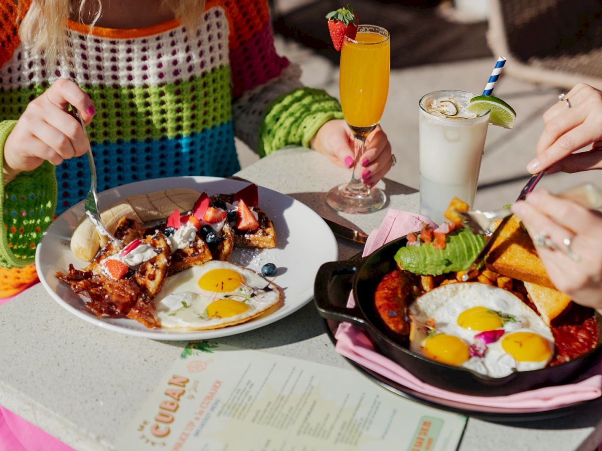 Two people enjoying a meal outdoors with plates of breakfast food, including eggs, waffles, and beverages on a table with a menu.