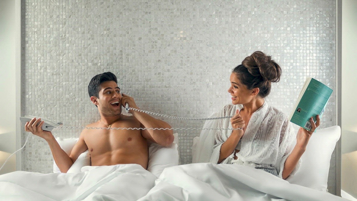 A couple is in bed; the man is shirtless on the phone, and the woman in a robe is smiling, holding a book and playfully holding the phone cord.