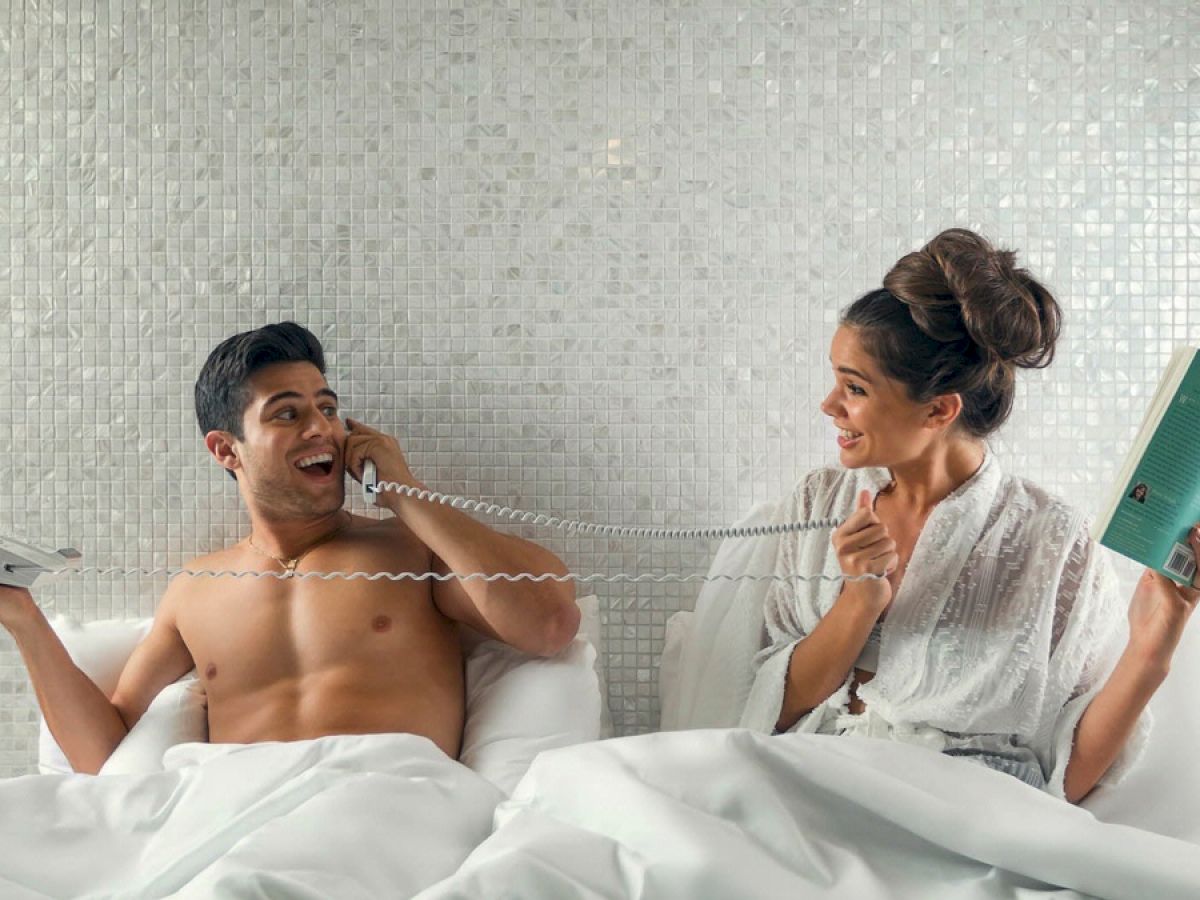 A couple is in bed; the man is shirtless on the phone, and the woman in a robe is smiling, holding a book and playfully holding the phone cord.