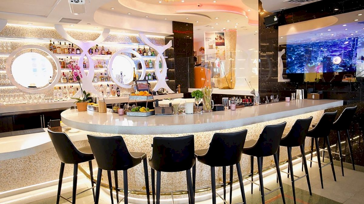 A modern bar with a curved countertop, black stools, illuminated shelves with bottles, and a large screen on the wall, in a stylish setting.