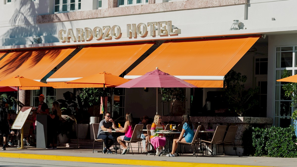 People are sitting at a sidewalk cafe outside the Cardozo Hotel with bright orange and pink umbrellas.