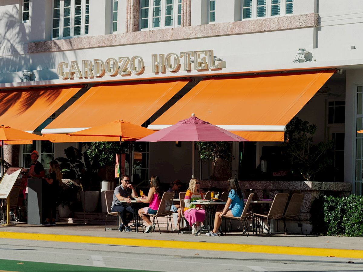 People are sitting at a sidewalk cafe outside the Cardozo Hotel with bright orange and pink umbrellas.
