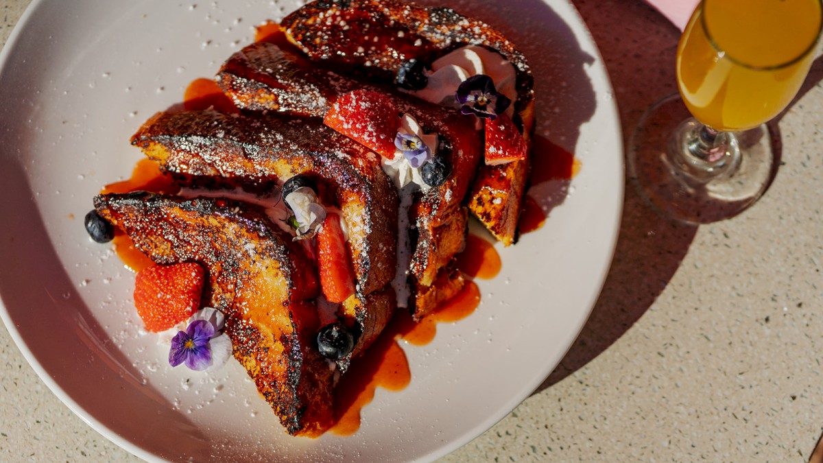 This image shows a plate of French toast topped with strawberries, blueberries, whipped cream, powdered sugar, and a garnish, next to a yellow drink.