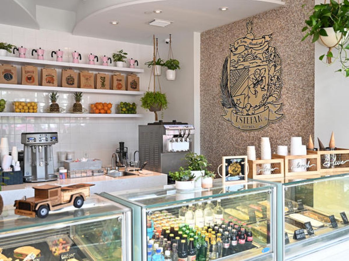 A modern and bright café interior with a counter displaying pastries, drinks, and a stylish coffee machine, alongside decor with plants and wall art.