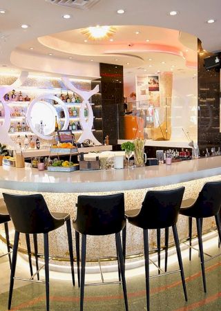 A modern, stylish bar with a sleek curved counter, high black chairs, and illuminated shelves stocked with bottles, featuring large TV screens.