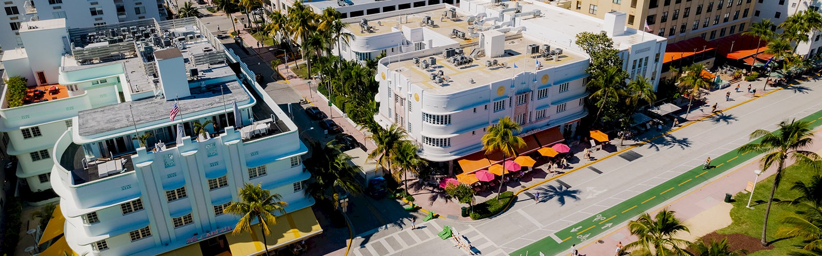 An aerial view of a cityscape with palm trees, buildings, a bike lane, and sidewalks, under a clear blue sky.