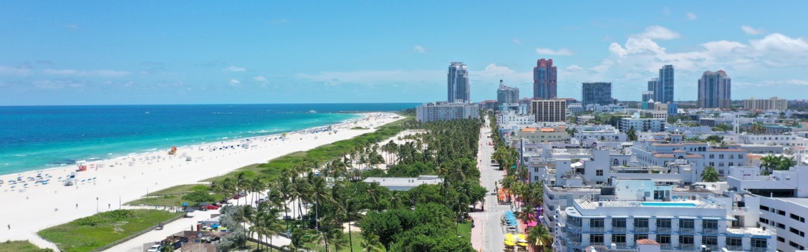 A coastal cityscape with a sandy beach, green space, and adjacent urban architecture, set against a blue sky with scattered clouds.