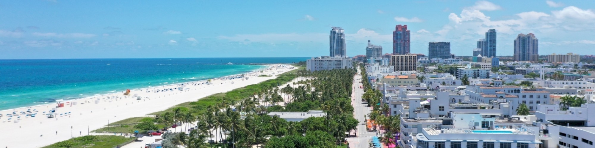 A coastal cityscape with a sandy beach, green space, and adjacent urban architecture, set against a blue sky with scattered clouds.