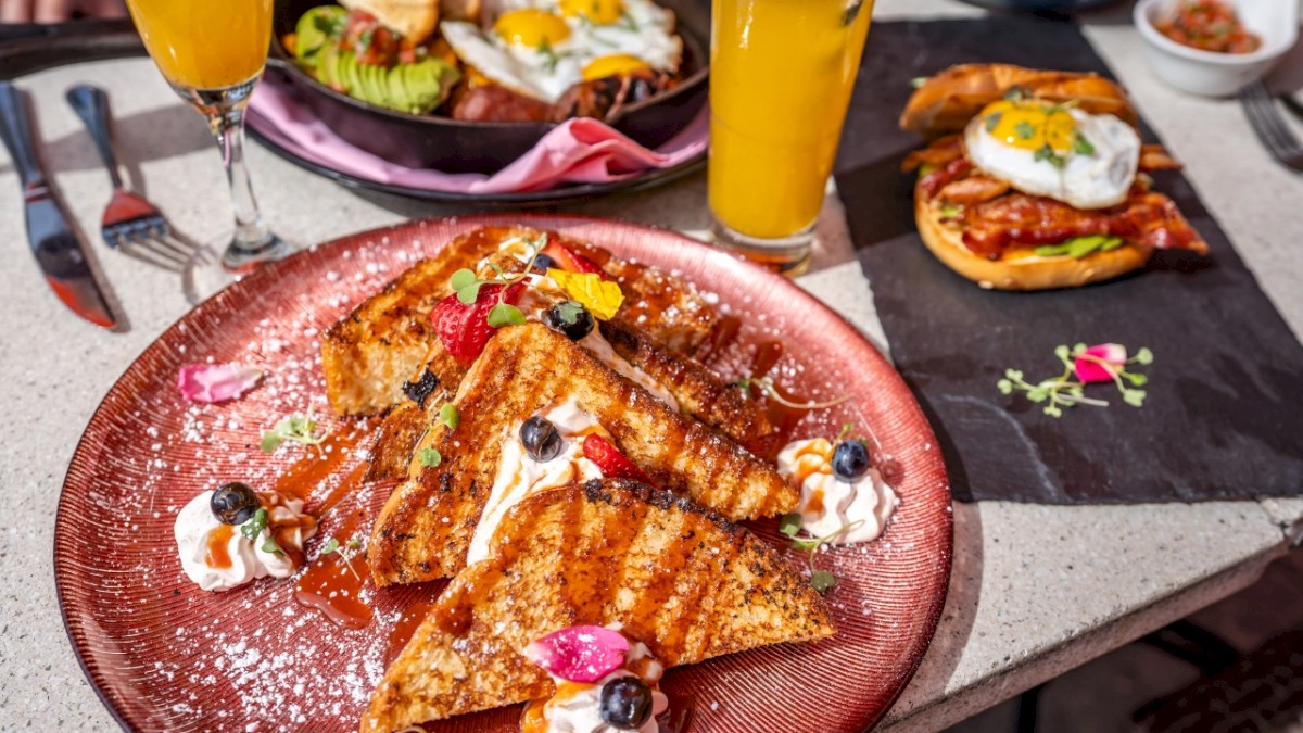 French toast with fruit and whipped cream, mimosas, a dish with avocado toast and eggs, and toast with bacon and eggs on a table.