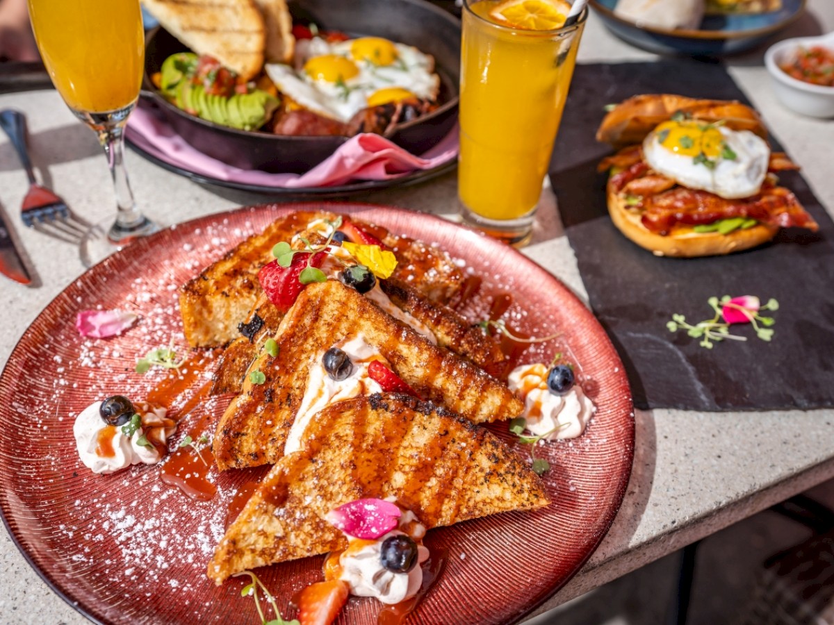 French toast with fruit and whipped cream, mimosas, a dish with avocado toast and eggs, and toast with bacon and eggs on a table.