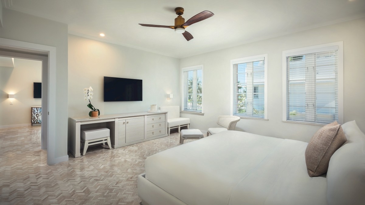 A modern bedroom with light decor featuring a bed, a TV on a stand, a ceiling fan, and windows allowing natural light to fill the room.