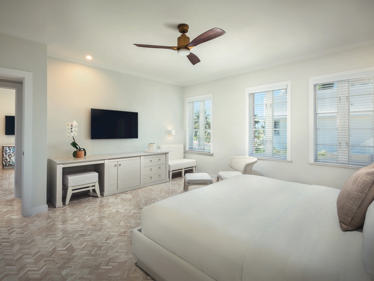 A modern bedroom with light decor featuring a bed, a TV on a stand, a ceiling fan, and windows allowing natural light to fill the room.