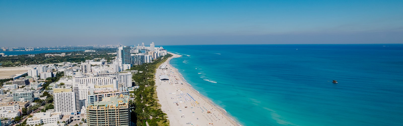 Aerial view of a coastal city with high-rise buildings along a sandy beach, clear turquoise water, and a bright blue sky.