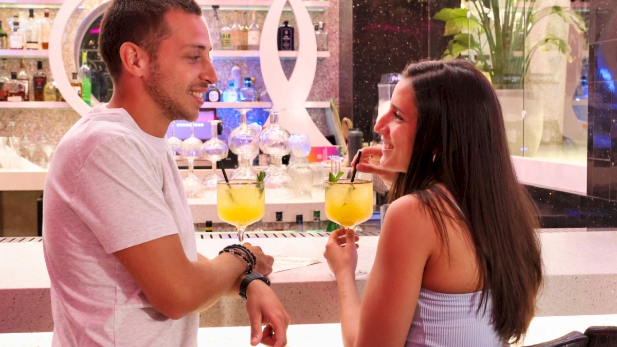 A man and a woman are sitting at a bar, smiling and holding yellow cocktails with straws, surrounded by bottles and plants.