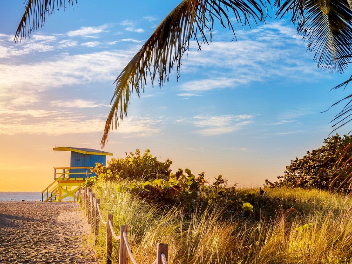 A picturesque beach with a lifeguard tower, sandy path, lush greenery, and palm trees under a vibrant sunrise or sunset sky.