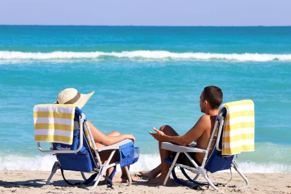 Two people sitting on beach chairs with yellow-striped towels, facing the ocean on a sunny day, engaged in conversation.