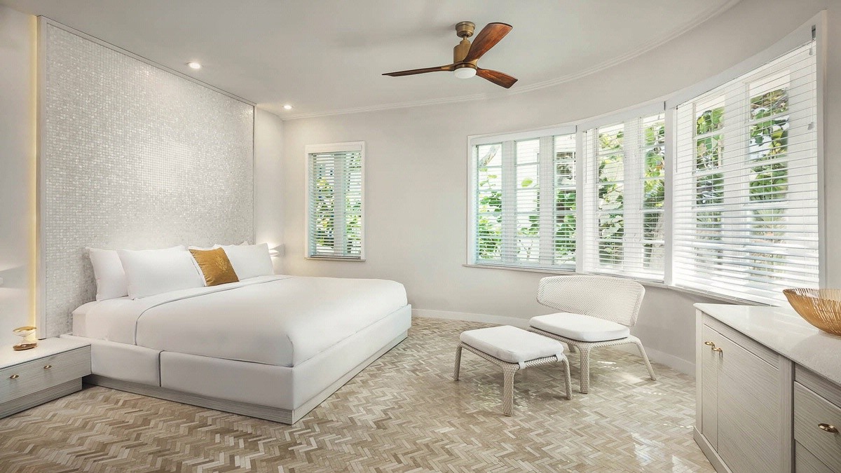 A bright, modern bedroom with a white bed, mosaic floor, white walls, a ceiling fan, a chair with an ottoman, and large windows with blinds.