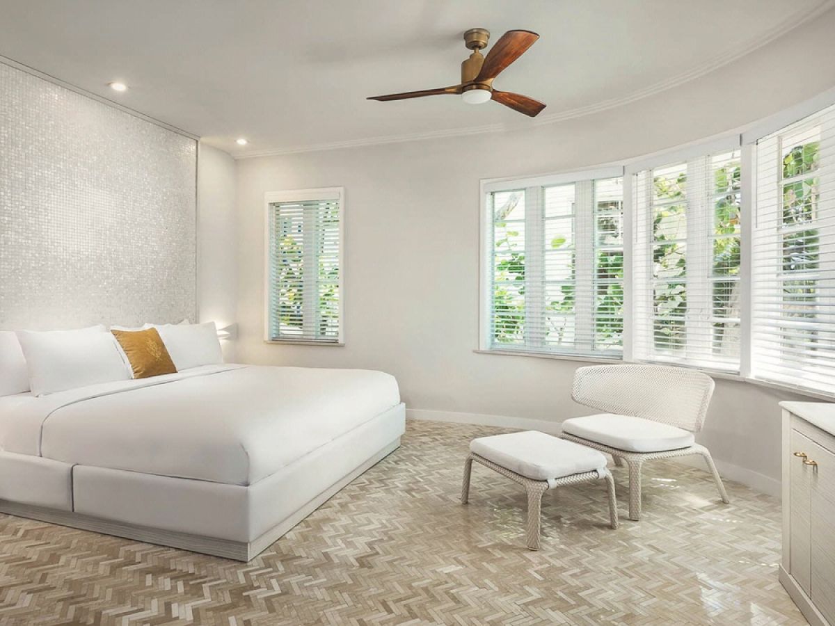 A bright, modern bedroom with a white bed, mosaic floor, white walls, a ceiling fan, a chair with an ottoman, and large windows with blinds.