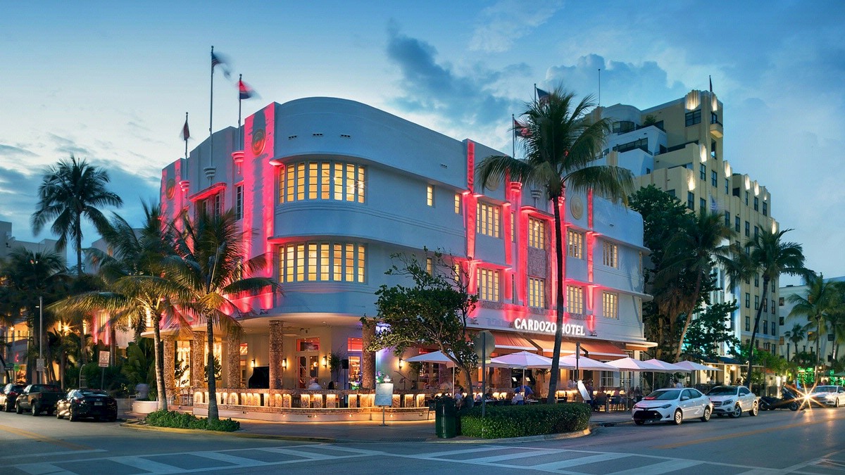 This image shows a brightly lit art deco building at dusk with palm trees and cars towards the side on a busy street corner.