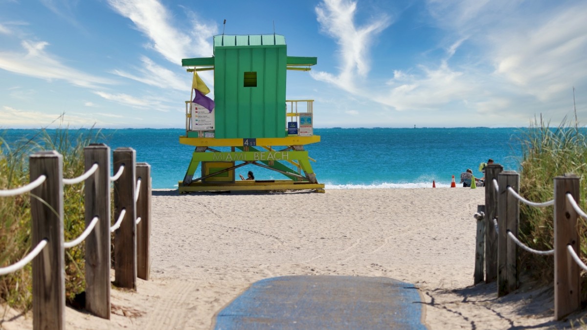 A colorful lifeguard tower on a sandy beach with a path leading to it, surrounded by blue skies and the ocean in the background.