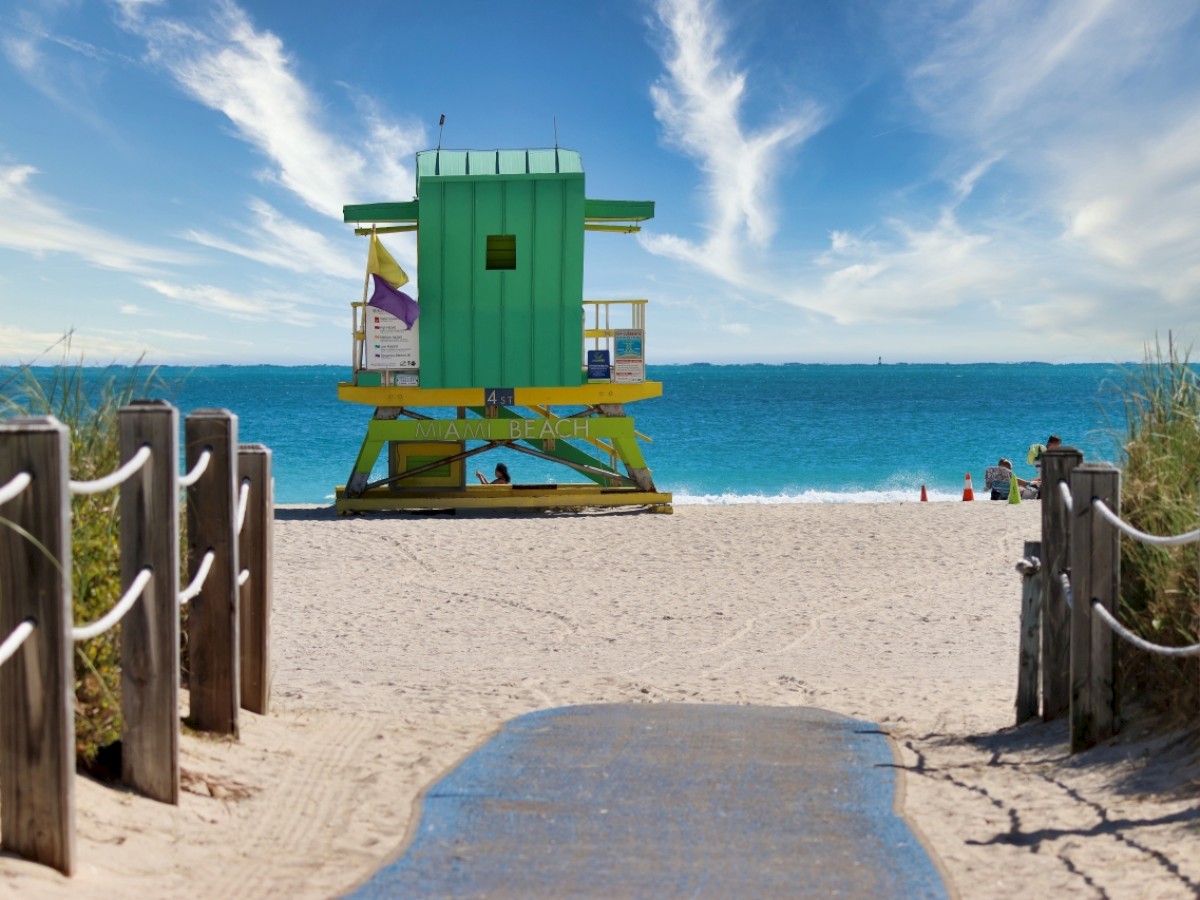 A colorful lifeguard tower on a sandy beach with a path leading to it, surrounded by blue skies and the ocean in the background.