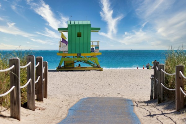 A beach scene with a colorful lifeguard tower, sandy pathway, wooden posts, and a bright blue sky with wispy clouds.
