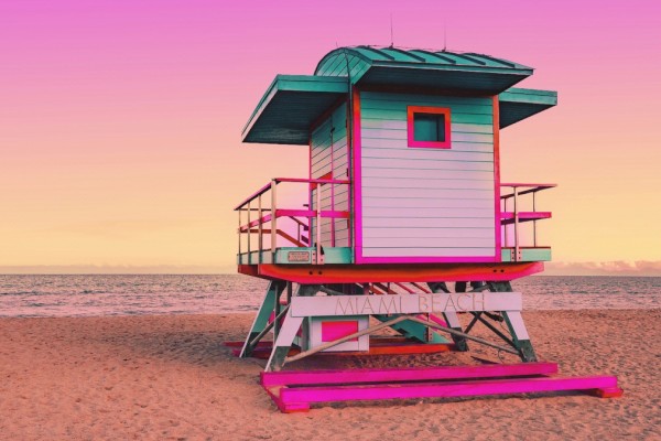 A colorful lifeguard tower stands on a sandy beach under a pink and orange sky at sunrise or sunset, with the ocean in the background.